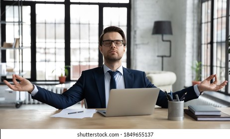 Front View Peaceful 30s Young Businessman Male Ceo Manager In Formal Wear Sitting Alone At Table With Folded In Mudra Gesture Hands, Doing Yoga Breathing Exercises, Reducing Stress At Workplace.