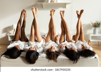 Front view playful happy young mixed race well groomed women best friends in silk gowns lying on comfortable bed with raised fit slim legs, enjoying spa weekend free relaxed time together in hotel.