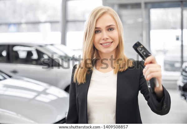 Front view of pleasant female car dealer in\
white shirt and black suit keeping car keys at hands in auto show\
room. Woman looking at camera, smiling and showing keys. Concept of\
car selling.