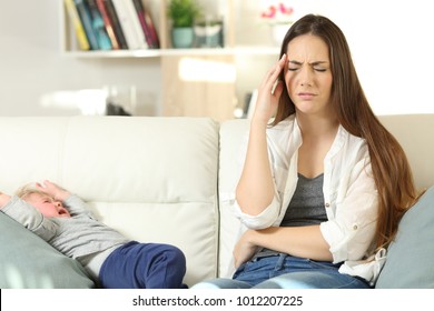 Front View Portrait Of An Annoyed Mother With Her Baby Son Crying Sitting On A Couch In The Living Room At Home