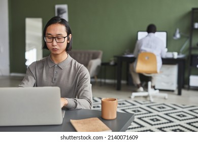 Front View Portrait Of Asian Man In Casual Wear Using Laptop While Working In Modern Office Against Green, Copy Space