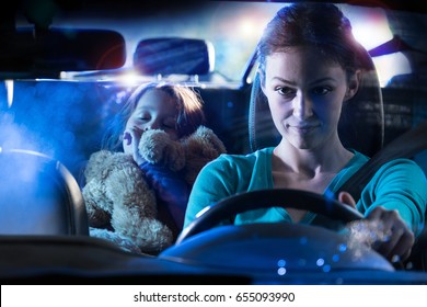 Front View. Portrait Of A Beautiful Woman Driving Her Car At Night In The Rain. In The Back Her Little Girl Fell Asleep Her Teddy In The Arms