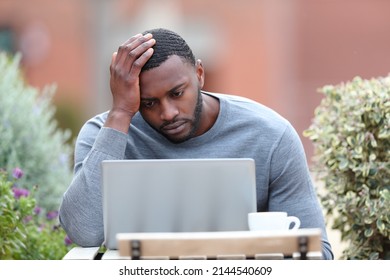 Front view portrait of a concerned man with black skin using laptop in a bar terrace
