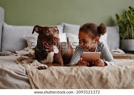 Front view portrait of cute African-American girl lying on bed with big pet dog and smiling, copy space