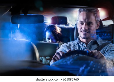 Front View. Portrait Of A Handsome Man Driving His Car At Night In The Rain. At The Back His Daughter Plays With A Digital Tablet