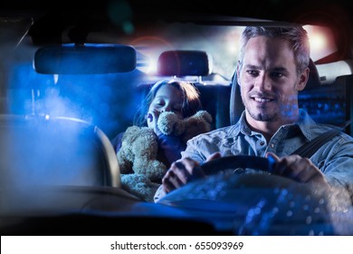 Front View. Portrait Of A Handsome Man Driving His Car At Night In The Rain. In The Back His Little Girl Fell Asleep Her Teddy In The Arms