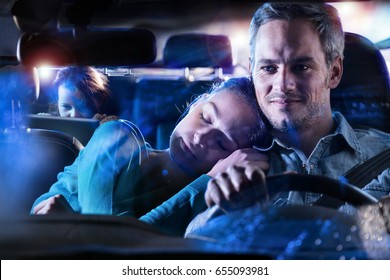 Front View. Portrait Of A Handsome Man Driving His Car At Night In The Rain, His Wife Fell Asleep On His Shoulder. At The Back Their Daughter Plays With A Digital Tablet