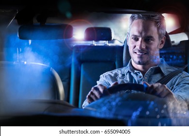 Front View. Portrait Of A Handsome Man Driving His Car At Night In The Rain. The Traffic Lights Behind Him