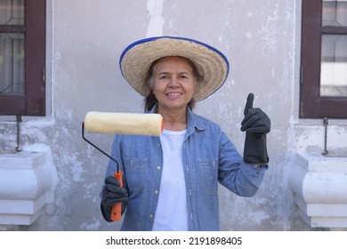 Front View, Portrait Of A Happy Asian Female Construction Worker Wearing Hay Hat And Gloves, Holding Paint Roller, Smiling, Putting Thumb Up. Home Renovation, Retiree Lifestyle Concept.