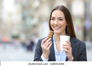 Front View Portrait Of A Happy Executive Woman Holding Cereal Snack Bar And Takeaway Coffee In The Street