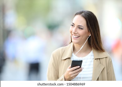 Front view portrait of a happy woman wearing earbuds listening to music on smart phone looking at side in the street - Powered by Shutterstock