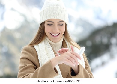 Front view portrait of a happy woman applying moisturizer cream to hydrate hands with a snowy mountain in the background