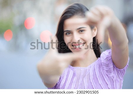 Front view portrait of a happy woman framing with hands looking at you in the street