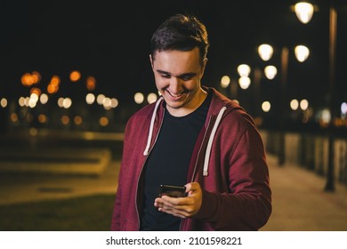 Front view portrait of one young adult caucasian man photographer standing outdoor in the city or town at night holding using mobile phone to read or send text message sms browse internet happy smile - Powered by Shutterstock