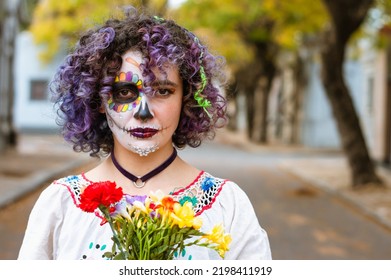 front view portrait of serious young woman with curlers holding flowers and with artistic makeup representing La Calavera Catrina, standing outdoors looking at camera with copy space.