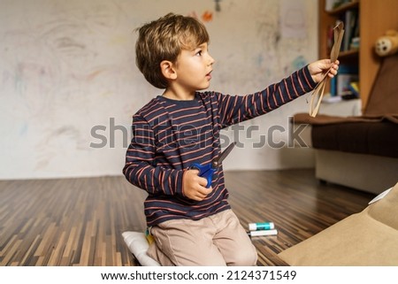 Front view portrait of small boy four years old child sitting on the floor at home alone holding scissors looking to the side - childhood development and leisure concept