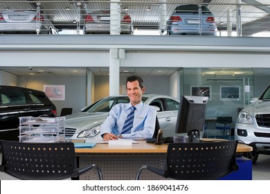 Front view portrait of a smiling car salesman sitting at the desk in the large car showroom. - Powered by Shutterstock