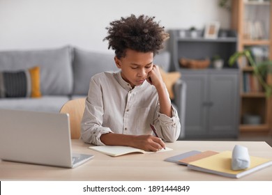 Front view portrait of teenage African-American boy doing homework and writing in notebook while sitting at desk with laptop, copy space
