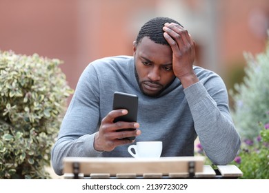 Front view portrait of a worried man with black skin checking phone in a coffee shop
