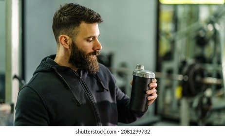 Front View Portrait Of Young Caucasian Man Athlete In Black Hoodie Male Standing In The Gym Holding Protein Supplement Shaker Supplementation In Training Waist Up Black Hair And Beard Copy Space Drink