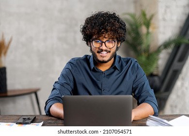 Front View Of Positive Hindi Man In Smart Casual Shirt Using Laptop While Sitting At The Desk In His Flat. Young Indian Male Student Watching Webinars, Educational Courses, Learning On The Distance