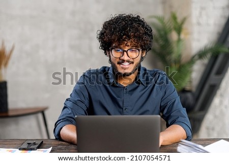 Front view of positive Hindi man in smart casual shirt using laptop while sitting at the desk in his flat. Young Indian male student watching webinars, educational courses, learning on the distance