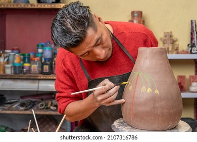 Front view potter using a paintbrush to decorate with drawings a clay pot he has just created in his studio.