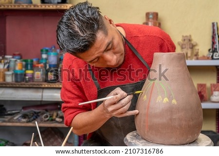 Front view potter using a paintbrush to decorate with drawings a clay pot he has just created in his studio.