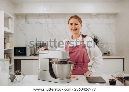 Front view of pretty joyful young woman, professional confectioner in white uniform and hat, using an electric planetary mixer for whipping egg whites, pouring sugar into the bowl. 