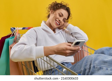 Front view of pretty young female sitting in cart with smartphone. Beautiful girl with curly hair holding mobile phone, looking, smiling, scrolling. Concept of modern life.