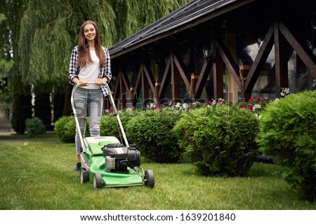 Front view of pretty young smiling woman using lawn mower on backyard, looking at camera. Female gardener working in summer, cutting grass in backyard. Concept of gardening, work, nature.