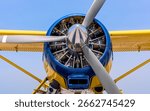 Front view of a radial engine on a floatplane with a vivid yellow and blue color scheme. The reflective spinner and propeller blades dominate the frame, with detailed mechanical components visible.