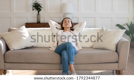 Front view relaxed young 30s woman sleeping on comfortable couch with folded hands behind head, enjoying weekend leisure lazy time, napping daydreaming or meditating alone in modern living room.