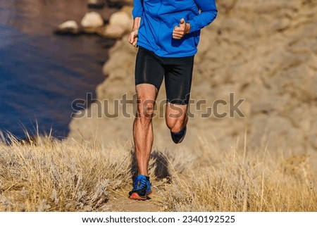 front view runner running mountain uphill in blue jacket and black tights, trail dry grass