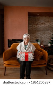 Front View Of Sad And Bored Elder Woman Sitting Alone On A Sofa With Wrapped Gift. No Family Visit Her On Christmas. Sadness During Coronavirus Pandemic. Covid Quarantine, Lockdown.