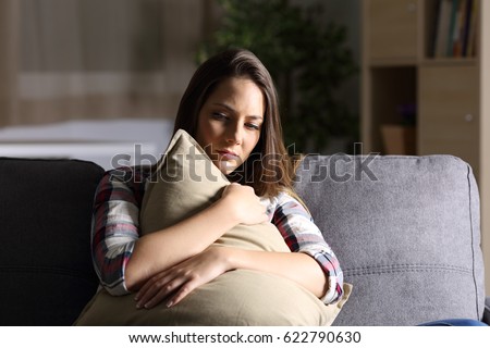 Front view of a sad single girl embracing a pillow sitting on a couch in the living room at home with a dark light in the background