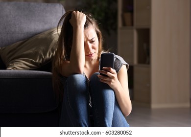 Front view of a sad teen checking phone sitting on the floor in the living room at home with a dark background
