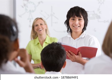 Front View Of A School Boy In Selective Focus Reading Out Of Test Booklet To Classmates Sitting In The Foreground And The�teacher Standing In The Background�