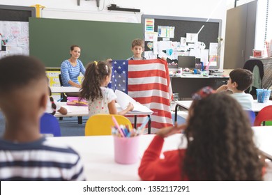 Front View Of A Schoolboy Holding An American Flag In Classroom At School With His Classmates In Foreground