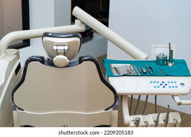 Front View Selective Focus Of Dental Instruments Set Placed On A Medical Disposable Blue Napkin On An Operator's Table Near A Blurred Empty Dental Chair With A Bright Dental Clinic Room In Background.