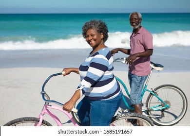 Front view of a senior African American couple standing on the beach with blue sky and sea in the background, leading their bicycles and smiling to camera - Powered by Shutterstock