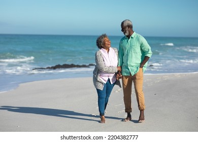 Front view of a senior African American couple walking on the beach with blue sky and sea in the background, holding hands and smiling at each other - Powered by Shutterstock
