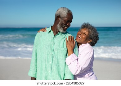 Front view of a senior African American couple standing on the beach with blue sky and sea in the background, embracing and smiling at each other - Powered by Shutterstock