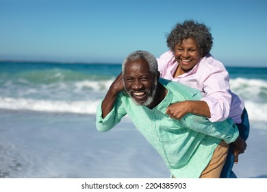 Front view of a senior African American couple standing on the beach with blue sky and sea in the background, piggybacking and smiling to camera - Powered by Shutterstock