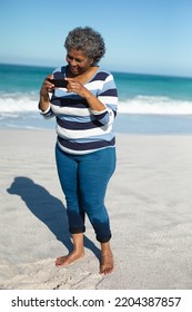 Front View Of A Senior African American Woman Standing On The Beach With Bare Feet, Blue Sky And Sea In The Background, Smiling And Using Her Mobile Phone