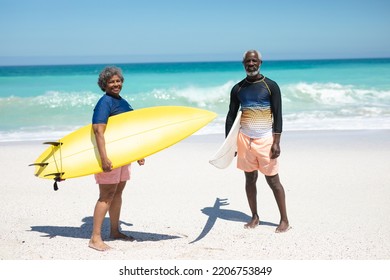 Front view of a senior African American couple on a beach in the sun, holding surfboards under their arms, looking to camera and smiling, with blue sky and calm sea in the background - Powered by Shutterstock