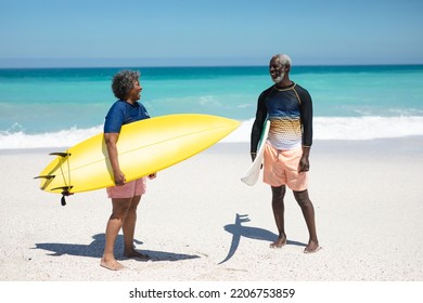 Front view of a senior African American couple on a beach in the sun, holding surfboards under their arms, looking at each other and smiling, with blue sky and calm sea in the background - Powered by Shutterstock