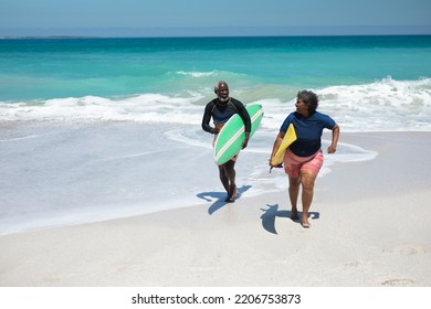 Front view of a senior African American couple on a beach in the sun, carrying surfboards under their arms and running, looking at each other and smiling, with blue sky and calm sea in the background - Powered by Shutterstock