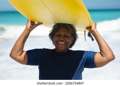 Front view of a senior African American woman on a beach in the sun, holding a surfboard on her head, looking to camera and smiling, with blue sky and calm sea in the background - Powered by Shutterstock