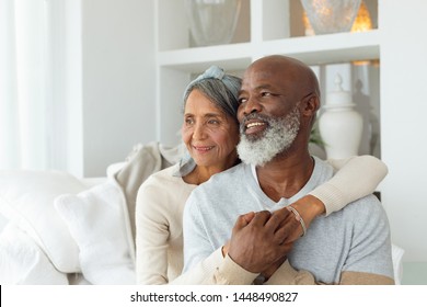 Vista frontal do casal sênior diversificado sentado em um sofá branco em uma casa de praia. Conceito autêntico de vida aposentado sênior